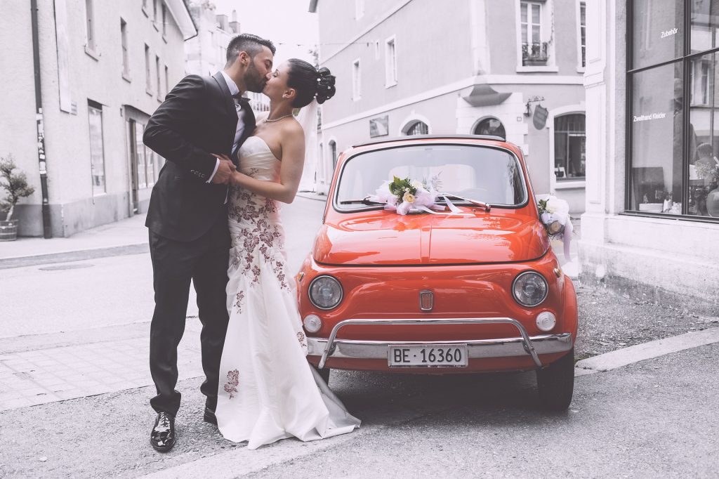 new wedding couple kissing at side of red car